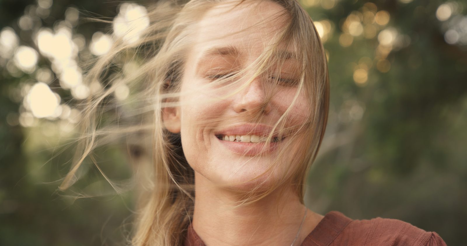 Woman with windblown hair, eyes closed, smiling, forest background
