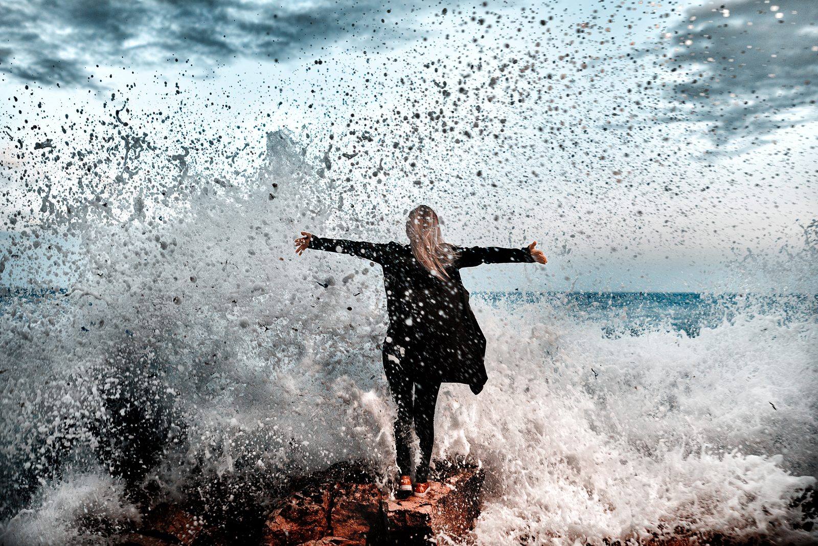 Woman with arms outstretched, standing on rocks as ocean waves crash and spray around her