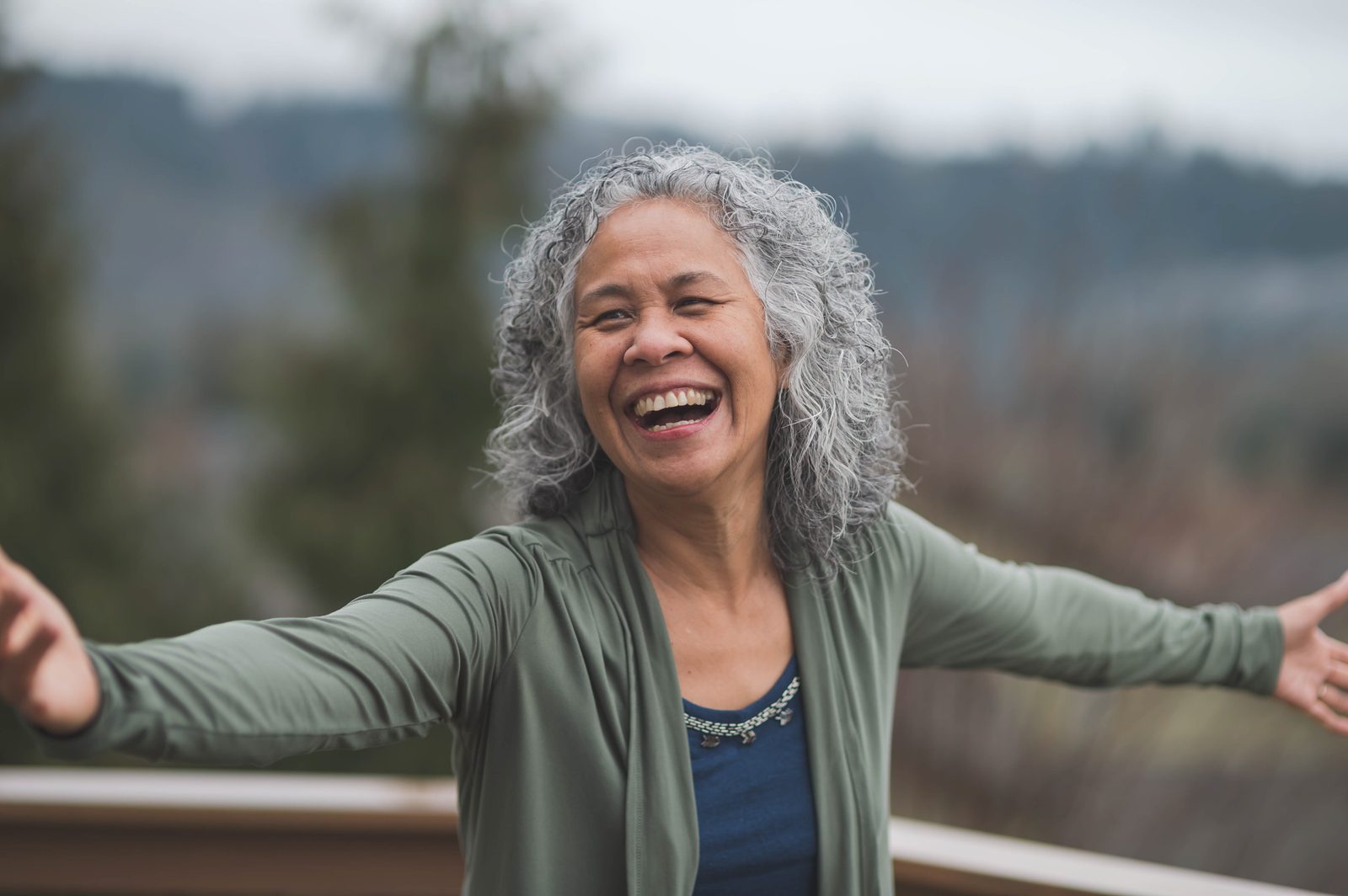 Older woman with curly gray hair, laughing with arms open wide, outdoors